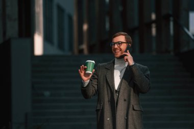 İnanılmaz güneş ışığı, elinde akıllı telefon ve bir bardak içki var. Resmi kıyafetli yakışıklı dışarıda, iş binasının yanında..