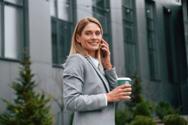 Yürümek, bir bardak içki içmek ve telefonla konuşmak. Resmi giysili kadın iş binasının yakınında dışarıda..