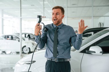 Gülümsüyor, şarj aletini tutuyor. Yakışıklı araba galerisi çalışanı elektrikli araçla çalışıyor..