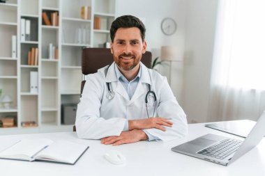 Masanın başında oturuyor. Resmi kıyafetli doktor dolapta çalışıyor..