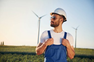 Mavi üniformalı ve güneş gözlüklü. Servis mühendisi yel değirmenleriyle sahada..