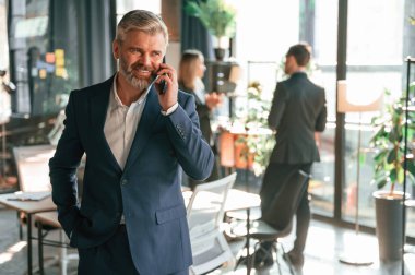 Adam ayakta ve telefonla konuşuyor. Bir grup iş adamı modern ofiste çalışıyor..
