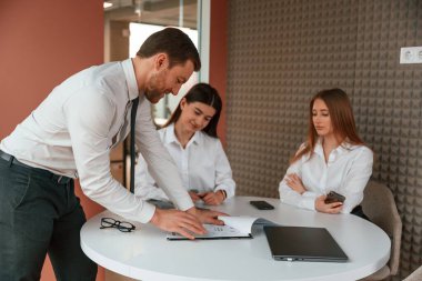Adam konuşuyor, belgeleri gösteriyor, projenin planını gösteriyor. Ofiste üç iş adamı birlikte çalışıyor..