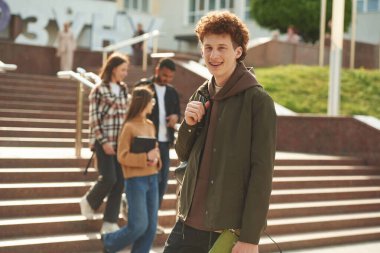 Kıvırcık saçlı adam arkadaşlarının önünde duruyor. Gündelik giysiler içinde dört genç dışarıda beraberler..