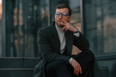 Sigara içmek, merdivenlerde oturmak. Resmi kıyafetli yakışıklı dışarıda, iş binasının yanında..