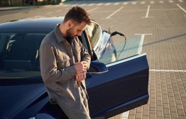 Otomobili şarj ediyorum. Adam dışarıda elektrikli arabasının yanında duruyor..