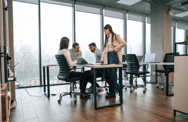 Masanın yanındaki büyük pencerelere yaslan. Ofiste birlikte çalışan dört kişi var..