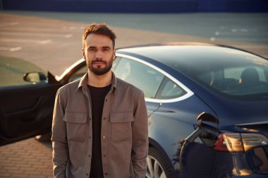 Muhteşem stil sahibi adam dışarıda elektrikli arabasının yanında duruyor..