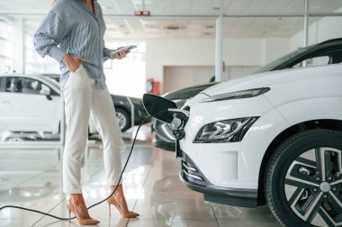 Ayakta duran, akıllı telefonu tutan, otomobil şarj oluyor. Elektrikli arabalı bir kadının yakın görüntüsü.