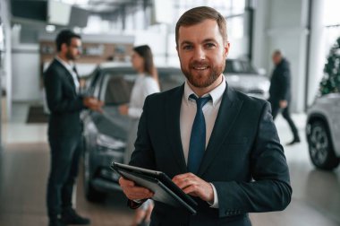 Güzel adam ayakta ve gülümsüyor. Araba galerisinde üç kişi birlikte çalışıyor..