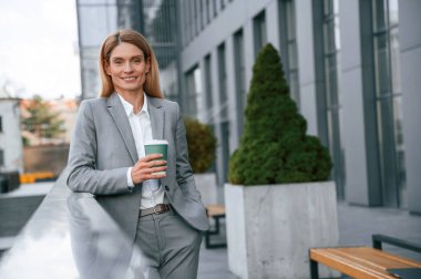 Gülümsüyor, elinde bir bardak içki tutuyordu. Resmi giysili kadın iş binasının yakınında dışarıda..