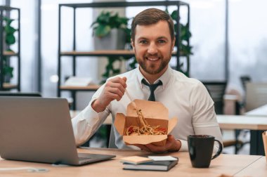 Dizüstü bilgisayarın yanında oturuyor. İş adamı resmi giysiler içinde ofiste ekolojik kutunun içinde udon yiyor..