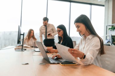 Masanın yanında oturuyor. Resmi kıyafetli iş adamları ofiste birlikte çalışıyorlar..
