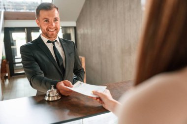Kadın ziyaretçiye hizmet ediyor. Otel resepsiyonunda siyah ceketli bir adam var..