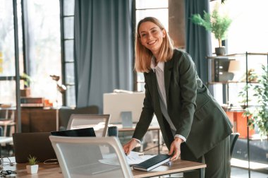 Ayakta ve gülümsüyor. Resmi giyinmiş genç bir bayan ofiste çalışıyor..