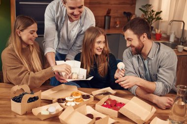Yiyecek karton eko kutularda. Arkadaşlar evde oturmuş, birlikte badem kurabiyesi, tatlı ve yaban mersini yiyorlar..