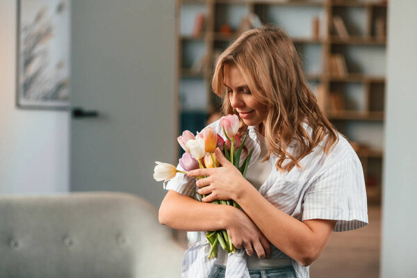 Holding the gift. Young woman is standing in domestic room with flowers.