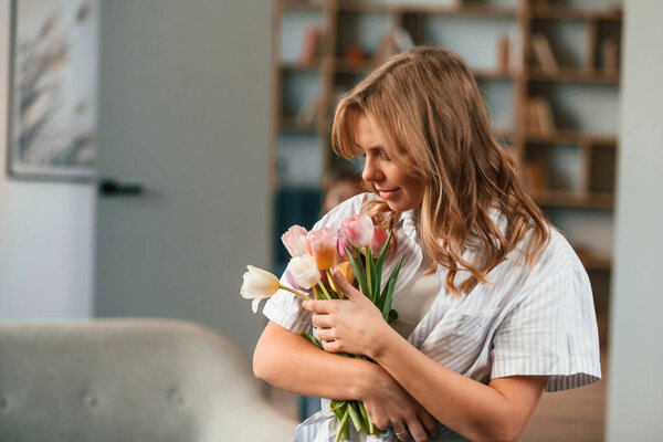 Holding the gift. Young woman is standing in domestic room with flowers.