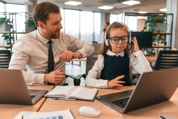 With adult man by the table with laptop. Little girl is in the office where people working together.