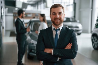 Yakışıklı bir adam iş arkadaşlarının önünde dikiliyor. Üç kişi araba galerisinde birlikte..