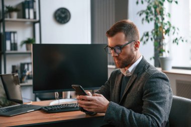 Oturmak, akıllı telefon kullanmak. Takım elbiseli iş adamı bilgisayarla ofiste..
