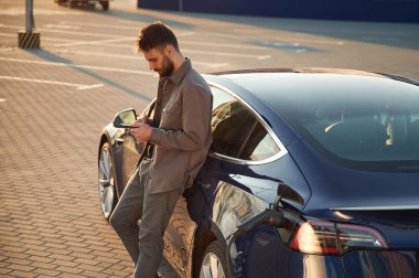 Arabaya yaslanıp elinde akıllı telefon tutarak. Şarj istasyonunda. Adam elektrikli arabasının yanında duruyor..