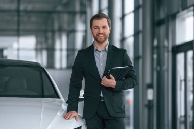 Elinde tabletle. Siyah takım elbiseli adam otomobilin yanındaki araba galerisinde..