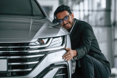 Ulaşımı kucaklıyorum. Takım elbiseli yakışıklı Hintli adam araba galerisinde araçla birlikte..