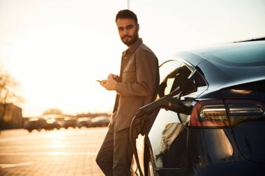 Arabaya yaslanıp elinde akıllı telefon tutarak. Şarj istasyonunda. Adam elektrikli arabasının yanında duruyor..