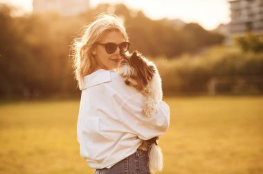 Güneş gözlüklü kadın yaz tarlasında duruyor ve kucağında küçük bir köpek var..