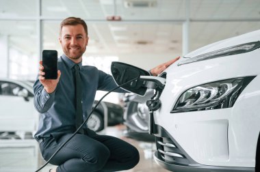 Oturmak, akıllı telefon göstermek, şarj etmek. Yakışıklı araba galerisi çalışanı elektrikli araçla çalışıyor..