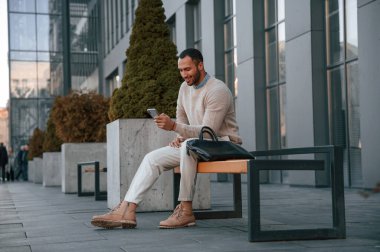 Akıllı telefon ve çantayla bankta. Yakışıklı siyah adam iş binasının yakınında dışarıda..