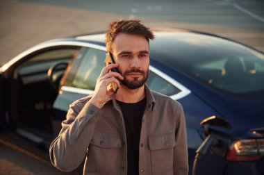 Otomobil şarj olurken arama yapmak. Adam dışarıda elektrikli arabasının yanında duruyor..