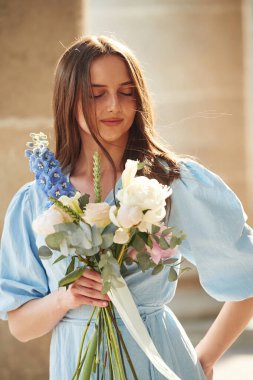 Beton sütunların yanında duruyordu. Güzel bir kadın elinde çiçeklerle dışarıda bekliyor..