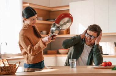 Kadın tavayı tutuyor ve bir adama vurmak istiyor. Genç çift mutfakta birlikte..