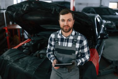 Elinde tabletle. Üniformalı adam araba galerisinde çalışıyor..