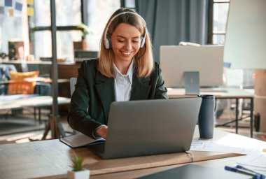 Gülümsüyor, dizüstü bilgisayar kullanıyor. Resmi giyinmiş genç bir bayan ofiste çalışıyor..
