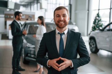 Güzel adam ayakta ve gülümsüyor. Araba galerisinde üç kişi birlikte çalışıyor..