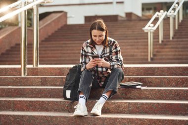 Akıllı telefon ellerde. Genç kız öğrenci üniversitenin merdivenlerinde oturuyor..