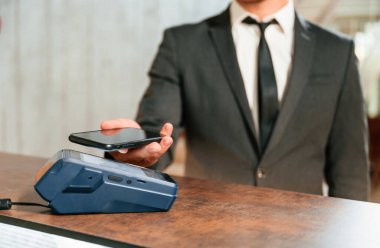 Ödemeyi telefon ve terminal kullanarak yapıyor. Otel resepsiyonunda siyah ceketli bir adam var..