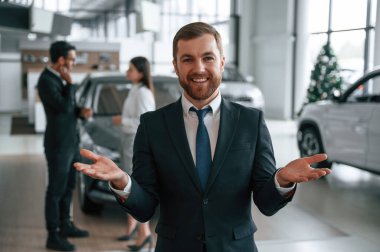 Yakışıklı bir adam iş arkadaşlarının önünde dikiliyor. Üç kişi araba galerisinde birlikte..