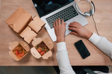 Dizüstü bilgisayarda daktilo eden bir adamın ellerinin üst görüntüsü. Karton eko kutulardaki yiyecekler masada..