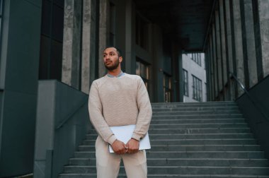 Ön manzara. Dizüstü bilgisayarın elinde duruyor. Yakışıklı siyah adam iş binasının yakınında dışarıda..