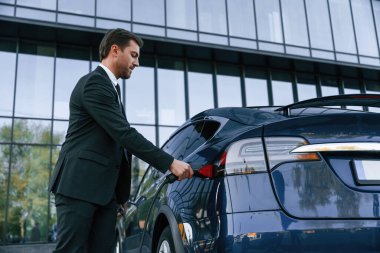 Hücum bekliyorum. İş adamı dışarıda elektrikli arabasının yanında duruyor..