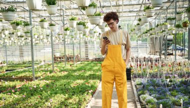 Akıllı telefon ellerde. Kıvırcık saçlı ve gözlüklü genç adam serada..