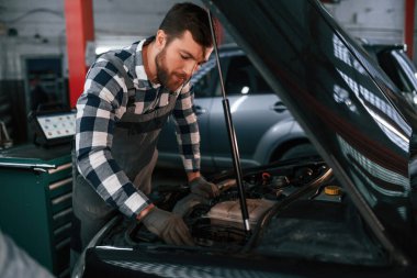 Siyah araba tamir ediliyor. Kaput açıldı. Üniformalı adam araba galerisinde çalışıyor..