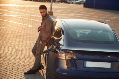 Şarj istasyonunda. Adam dışarıda elektrikli arabasının yanında duruyor..