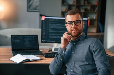 Yazılım geliştiricisi oturuyor ve poz veriyor. Resmi kıyafetli adam modern ofiste çalışıyor. Bilgisayar kullanılıyor.