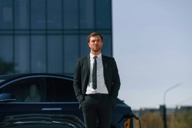 Ciddi bir yüz ifadesi. İş adamı dışarıda elektrikli arabasının yanında duruyor..