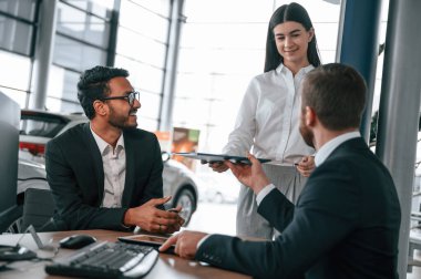 Kadın belgeleri veriyor. Araba galerisinde üç kişi birlikte çalışıyor..